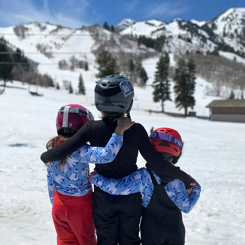 Three children in ski gear standing on a snowy slope with mountains in the background